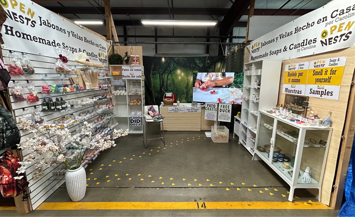 A retail display booth for 'OpenNests' handmade soaps and candles, featuring white shelving units, yellow and white signs, and a concrete floor with yellow markings inside a flea market.