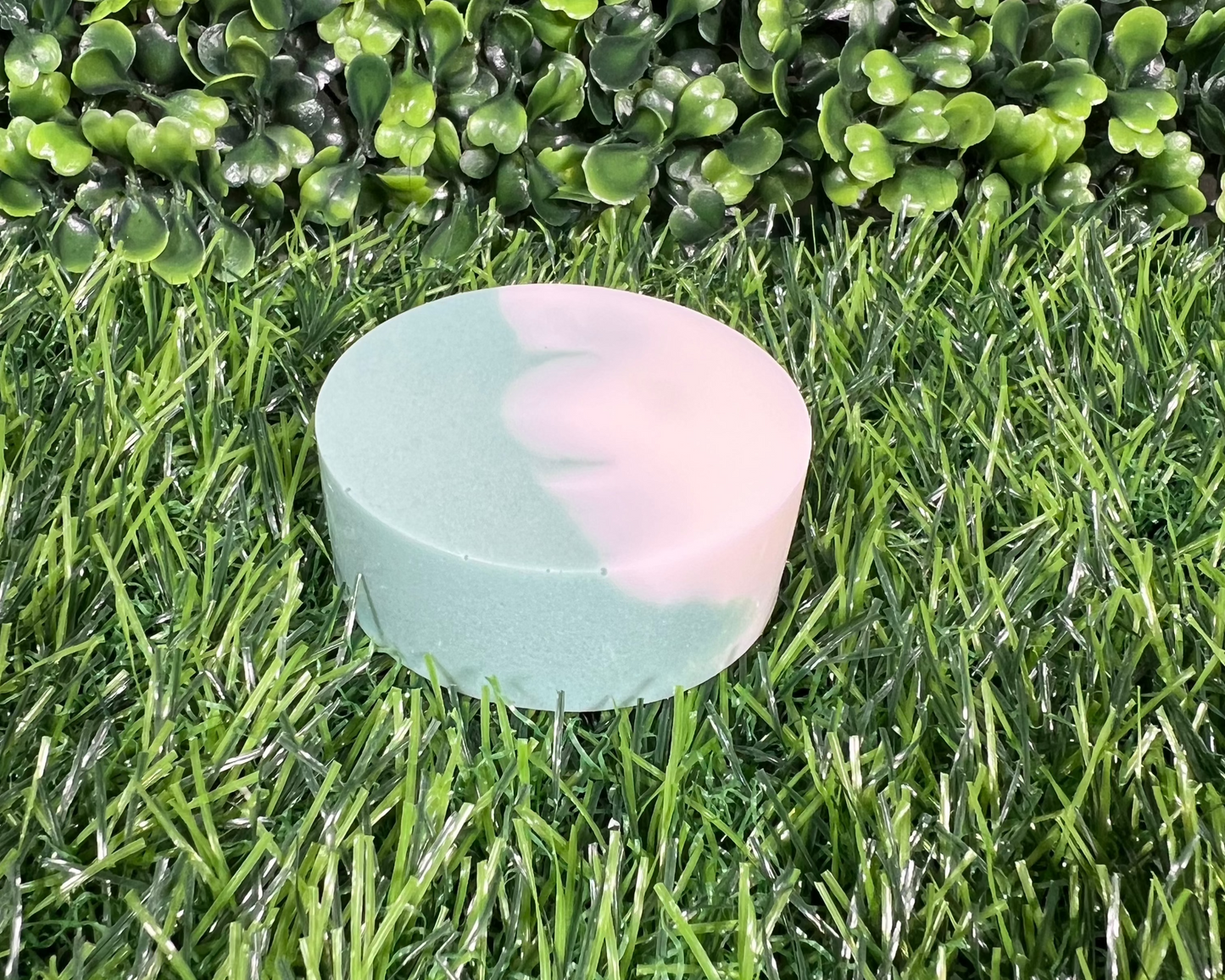 Circular shaped bar of mint and eucalyptus soap that is a mix of a green and white, displayed on a grass like surface.