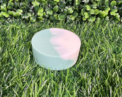 Circular shaped bar of mint and eucalyptus soap that is a mix of a green and white, displayed on a grass like surface.