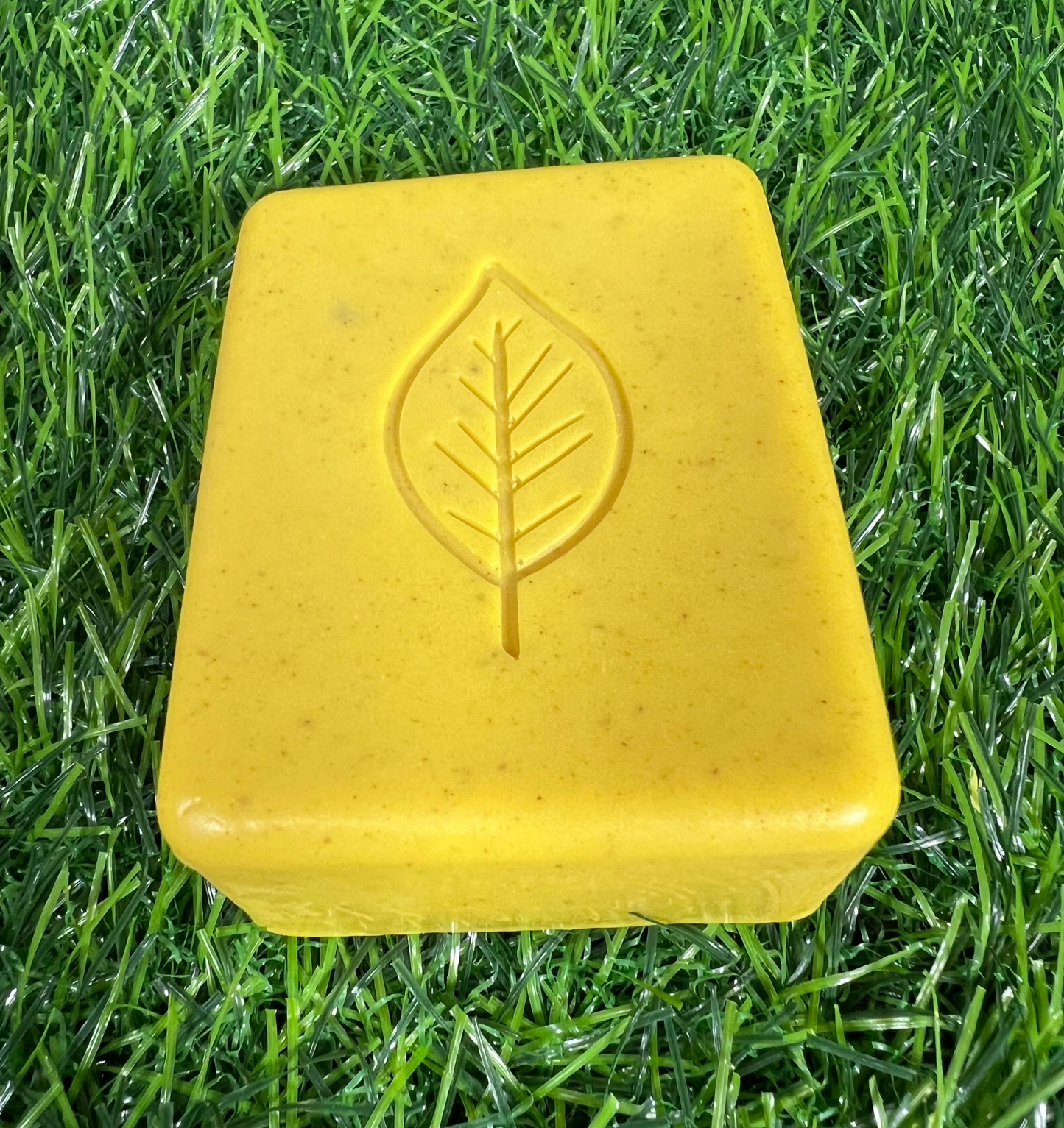 Rectangular turmeric yellow bar of soap with rounded edges and a leaf pattern on them, displayed on a green grass-like surface.