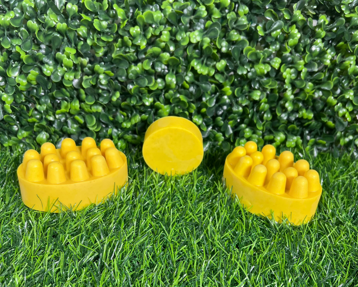 Three bars of turmeric soap with a yellow color and on a textured surface, displayed on a synthetic grass surface with a green leaf like background.