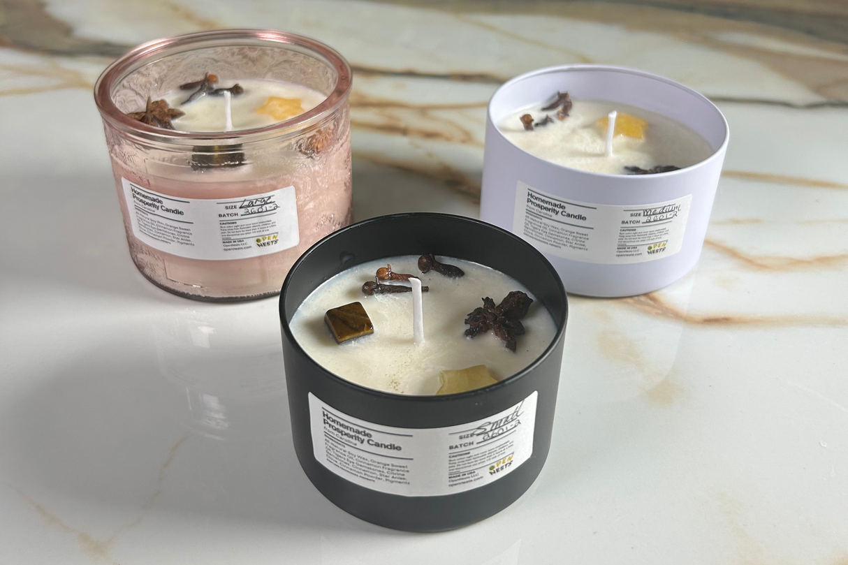 Three prosperity candles of varying sizes (2 metal tins (one black and one grayish white) and 1 pink glass jar)