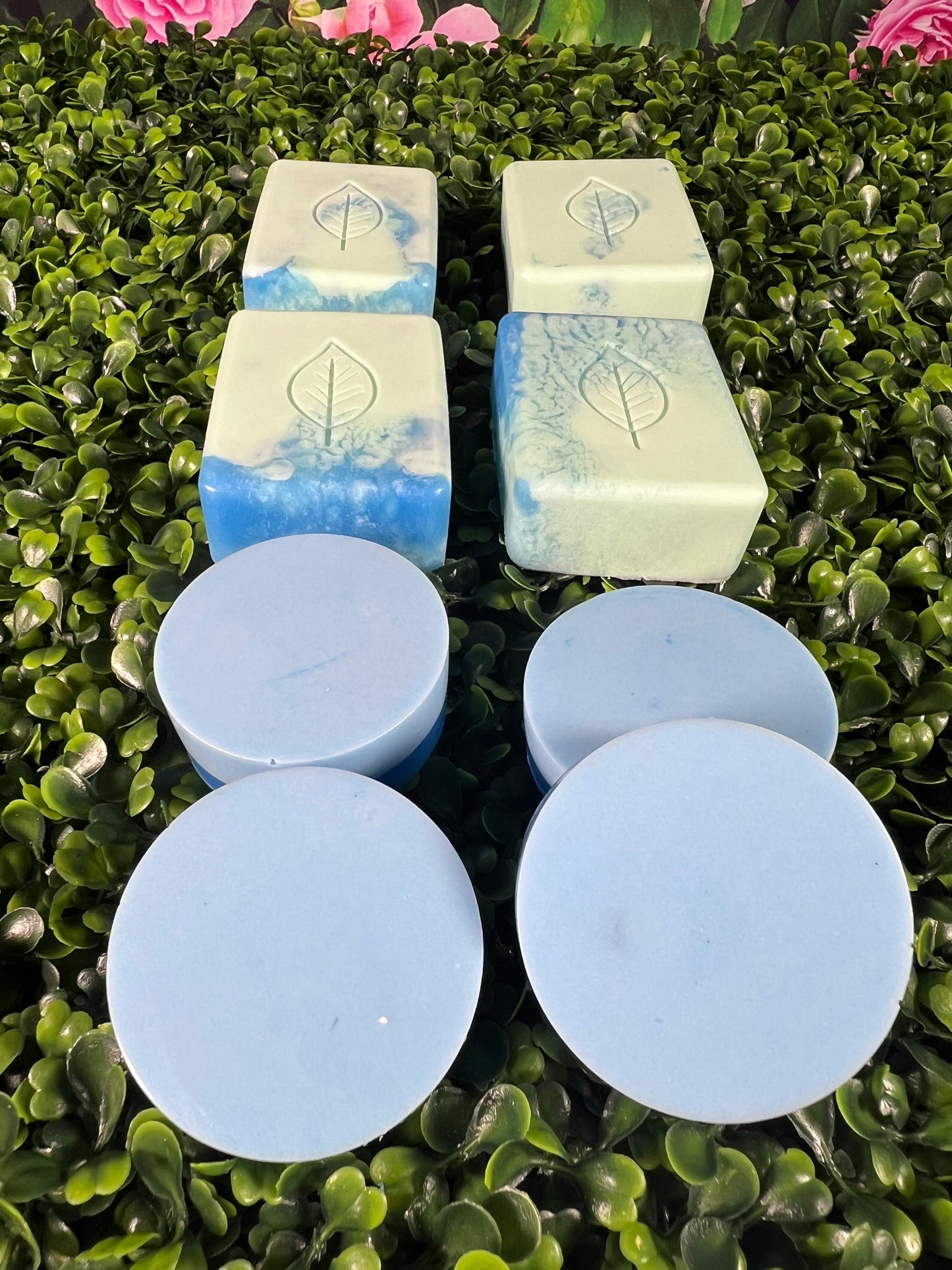 A variety of bar soaps with a blue and light green color scheme displayed on a green, leaf like surface, with some bars cut into circles and others in rectangular shapes.