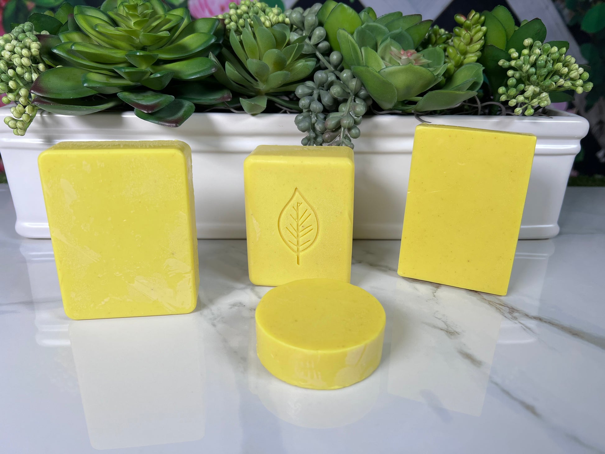 Multiple bars of homemade turmeric soap of varying sizes displayed on a surface with a white object in the background that resembles a soap dish, decorated with green plants.