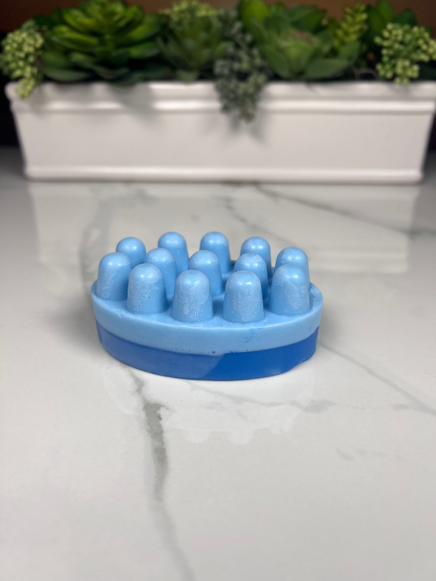 Scrub shaped bar of soap with dual blue color scheme (dark clear blue on the bottom layer and a light blue on the top layer where the scrub's nubs are) displayed on marble table.
