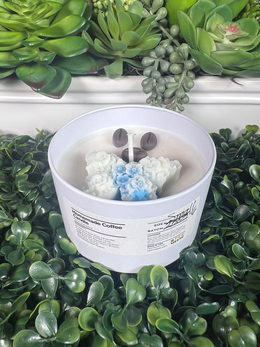 Candle with decorative elements (such as 3 wax coffee beans and a blue and white wax butterfly) in a white tin container on green foliage