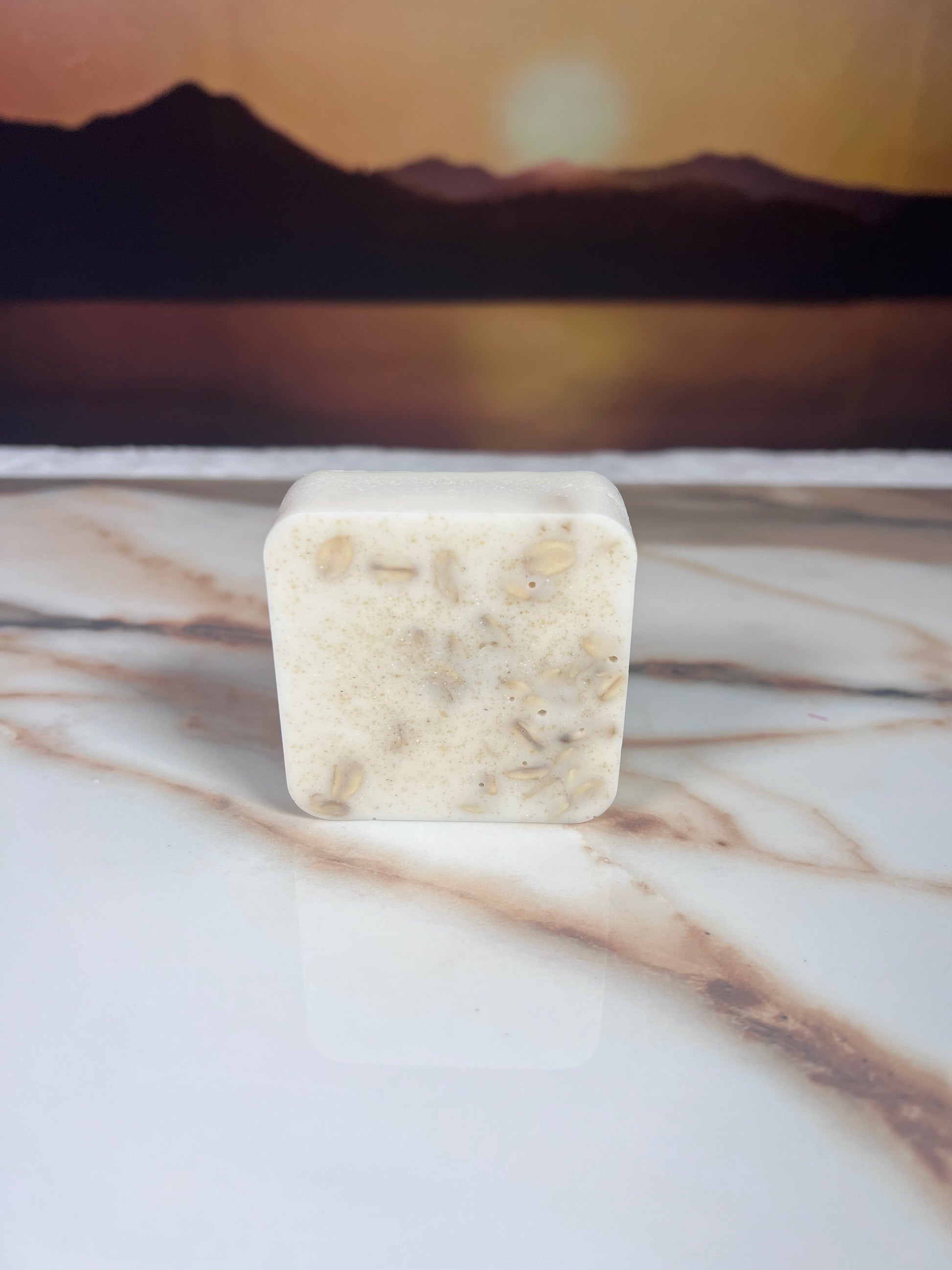 White square piece of soap with oatmeal pieces inside on top of a marble surface with a blurred mountainous background
